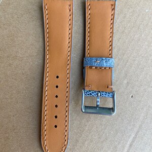 May include: A pair of watch straps with a light blue and gray snakeskin pattern. The straps are made of leather and have a tan color. The straps have stitching along the edges and a buckle.
