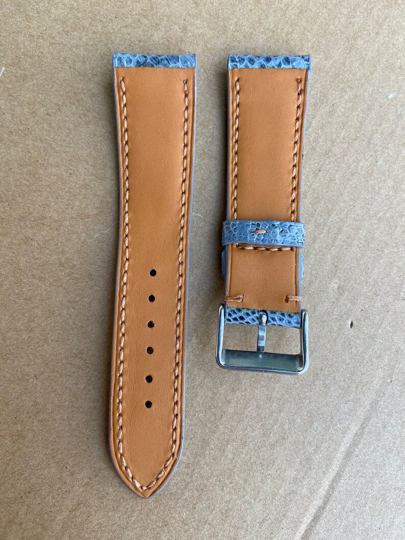 May include: A pair of watch straps with a light blue and gray snakeskin pattern. The straps are made of leather and have a tan color. The straps have stitching along the edges and a buckle.