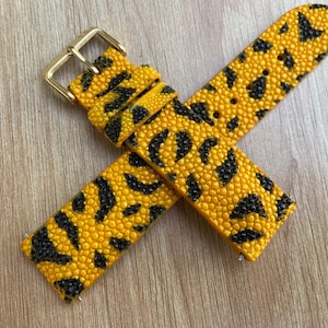 Puede incluir: Una correa de reloj de piel de raya con estampado amarillo y negro. La correa tiene una hebilla dorada.