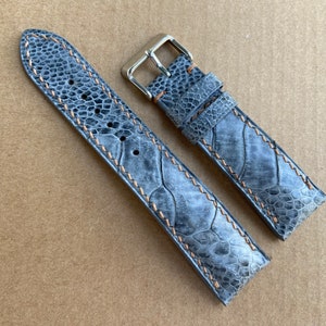 May include: Two gray leather watch straps with a light brown stitching detail. The straps are made of a textured material that resembles the skin of a reptile. The straps have silver metal buckles.