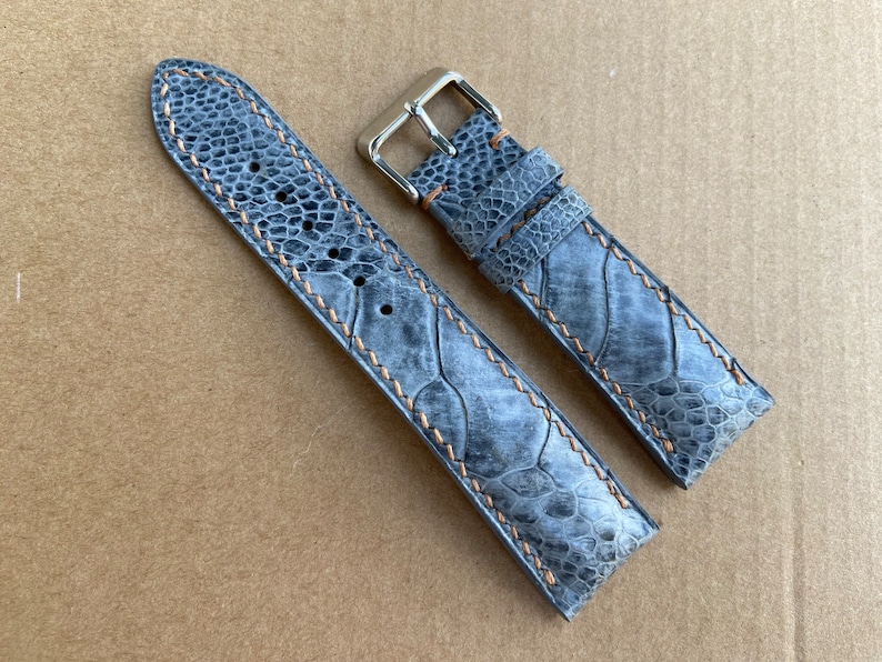 May include: Two gray leather watch straps with a light brown stitching detail. The straps are made of a textured material that resembles the skin of a reptile. The straps have silver metal buckles.