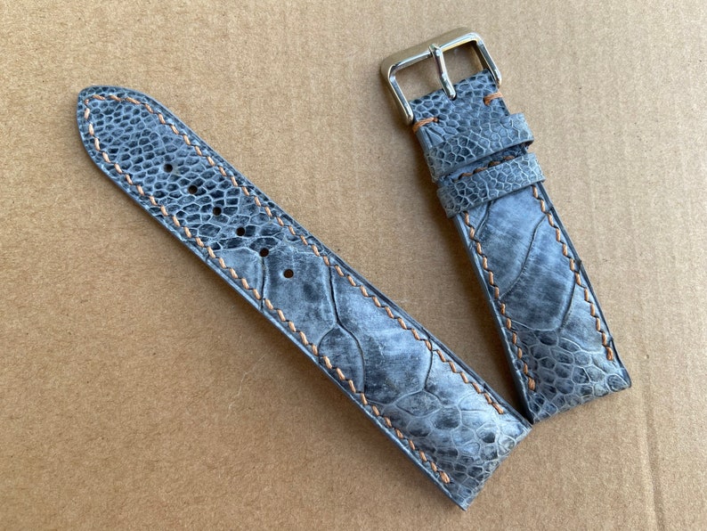 May include: A gray leather watch strap with a light brown stitching detail. The strap is made of a textured material that resembles a reptile skin.
