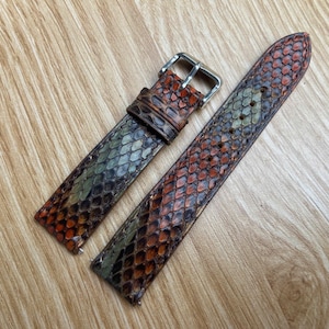 Puede incluir: Correa de reloj de dos piezas con un patrón de piel de serpiente en tonos marrón, naranja y verde. La correa tiene una hebilla plateada y está sobre una superficie de madera clara.