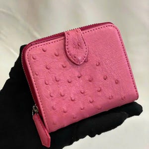 Könnte beinhalten: Ein pinkfarbenes, quadratisches Portemonnaie mit strukturierter Straußenlederoptik. Es hat einen Reißverschluss, einen Druckknopfverschluss und eine kleine Lasche. Das Portemonnaie wird von einer behandschuhten Hand gehalten. Ca. 10 cm x 10 cm.