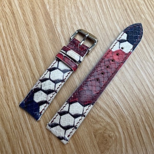 Op de afbeelding: Twee horlogebandjes met een reptielhuidpatroon in rood, wit en marineblauw. Eén bandje heeft een zilveren gesp. De bandjes hebben een geometrisch ontwerp en zijn geplaatst op een houten achtergrond.