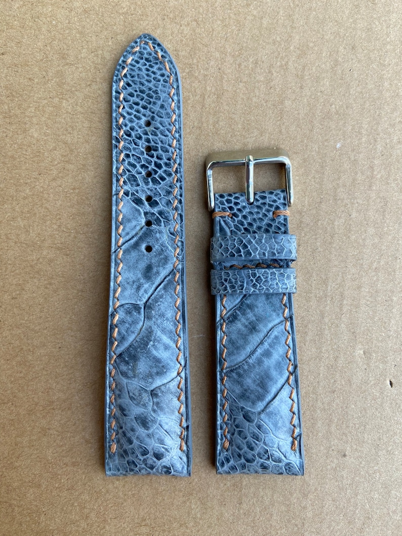 May include: A gray leather watch strap with a silver buckle and orange stitching. The strap is made of genuine ostrich leather.
