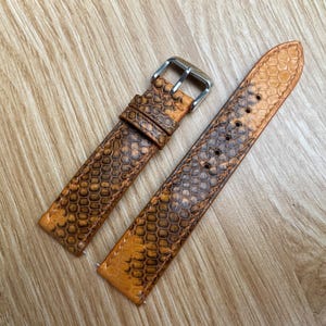 以下が含まれることがあります： 茶色とタン色の爬虫類革模様の2ピースの時計ストラップ。ストラップにはステンレス製のバックルが付いており、時計の文字盤に取り付けるように設計されています。革は質感のある外観をしています。