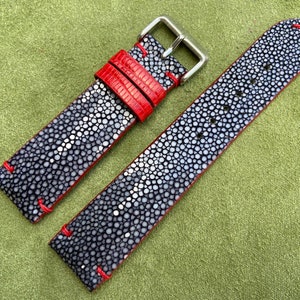 Puede incluir: Una correa de reloj de piel de raya gris y blanca con costuras rojas. La correa tiene una hebilla plateada.
