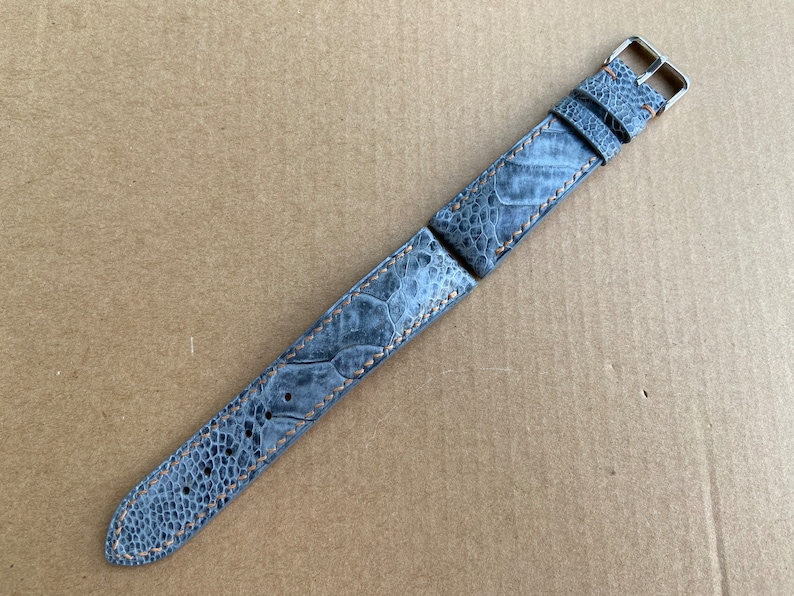 May include: A gray ostrich leather watch strap with orange stitching and silver hardware.