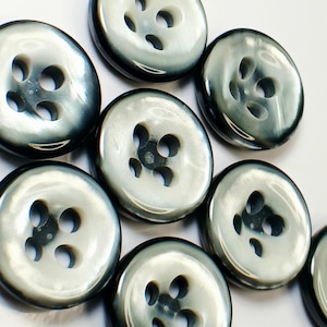 May include: A collection of pearlescent gray buttons with four holes each. The buttons have a slightly domed shape and a glossy finish, reflecting light. The buttons are arranged closely together, showcasing their design.