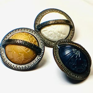 May include: Three antique buttons with intricate designs. One button features a tan globe design, another has a white carved relief, and the third is a dark blue oval with a carved pattern. Each button has a decorative metal rim.