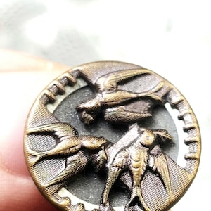 May include: A round, metallic button with a bronze-toned finish featuring a raised, embossed design of three birds in flight. The background of the button has a textured, dark gray surface, and the outer rim has a notched, gear-like edge.