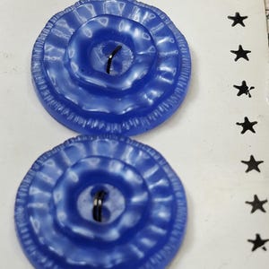 May include: Two round, bright blue buttons with a scalloped, ruffled edge and a central depression featuring two thread holes. The buttons are attached to a white card that displays the partial text New York and ED ARTISTS with black stars.