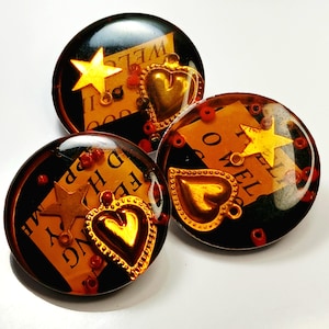 May include: Three round, black and gold resin buttons. Each button features a gold star, a gold heart, and a piece of paper with text. Red beads are scattered around the edges. The text on the paper reads "WELCOME".