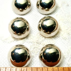May include: Six shiny, silver-colored, spherical beads arranged in two rows on a white lace surface. The beads reflect light, creating bright highlights. A wooden ruler is visible at the bottom of the image.