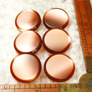 May include: Six round, copper-colored buttons with a glossy finish. The buttons have a lighter center and a darker, reddish-brown rim. A wooden ruler is partially visible in the image.