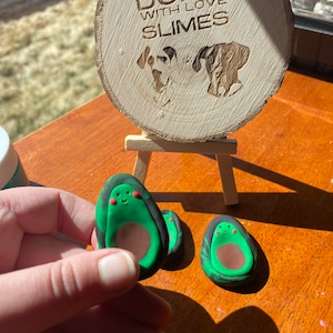 May include: Two green and black avocado-shaped clay ornaments with smiling faces. The ornaments are sitting on a wooden surface. The text "DUNN WITH LOVE SLIMES" is visible on a wooden sign in the background.