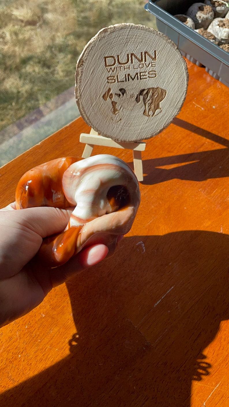 May include: A hand holds a brown and white slime with swirls. A wooden sign with the text "Dunn with love slimes" is in the background.
