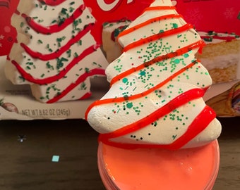 May include: A white frosted Christmas tree cookie with red and green sprinkles sits on top of a jar of pink slime. The cookie is topped with red icing and green sprinkles. The jar of slime is clear and has a red lid.