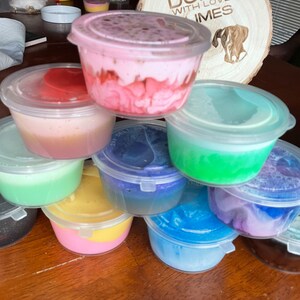 May include: A collection of small, clear plastic containers filled with colorful slime. The slime is in various shades of pink, blue, green, yellow, and purple. The containers are stacked on top of each other on a wooden surface.