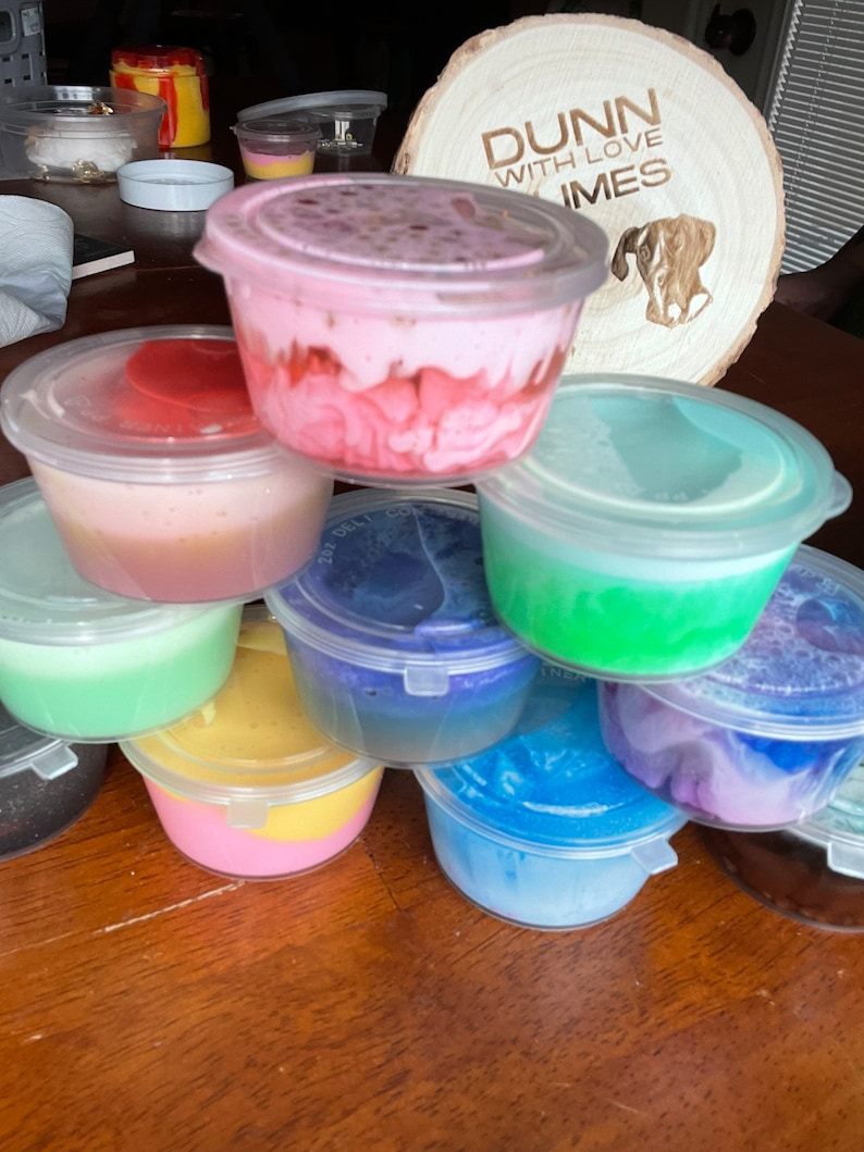May include: A collection of small, clear plastic containers filled with colorful slime. The slime is in various shades of pink, blue, green, yellow, and purple. The containers are stacked on top of each other on a wooden surface.