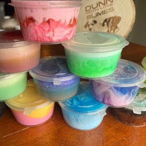 May include: A stack of small clear plastic containers filled with colorful slime. The containers are stacked in a pyramid shape. The slime is a variety of colors, including pink, green, blue, purple, and yellow. The containers are labeled "Dunn with Love Slimes".