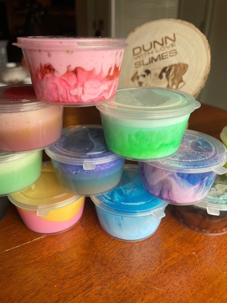 May include: A stack of small clear plastic containers filled with colorful slime. The containers are stacked in a pyramid shape. The slime is a variety of colors, including pink, green, blue, purple, and yellow. The containers are labeled "Dunn with Love Slimes".