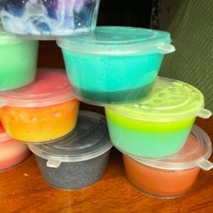 May include: A stack of seven small plastic containers filled with different colored slime. The slime is layered in some containers, with a lighter color on top of a darker color. The containers are stacked in a pyramid shape.