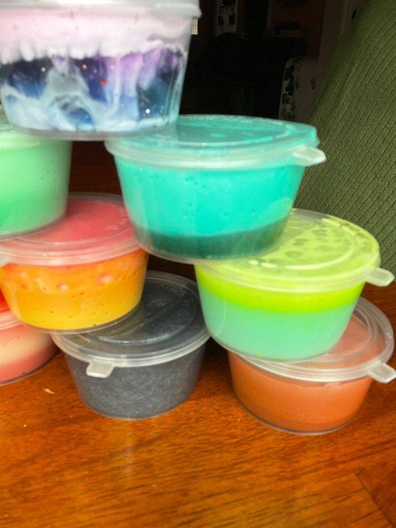 May include: A stack of seven small plastic containers filled with different colored slime. The slime is layered in some containers, with a lighter color on top of a darker color. The containers are stacked in a pyramid shape.