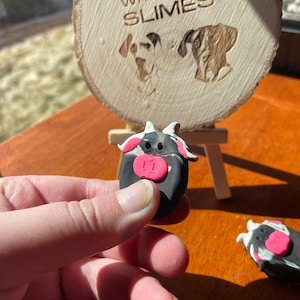 May include: A black and white cow figurine with a pink nose, made of polymer clay. The figurine is being held in a hand. The background shows a wooden sign with the text "Dunn with love slimes".