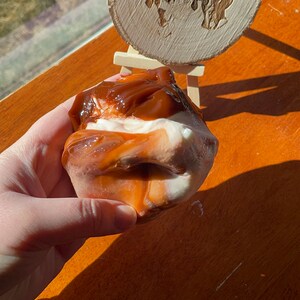 May include: A close-up of a brown and white slime with a caramel-colored swirl. The slime is being held in a hand. A wooden sign with the text "Dunn with Love Slimes" is in the background.