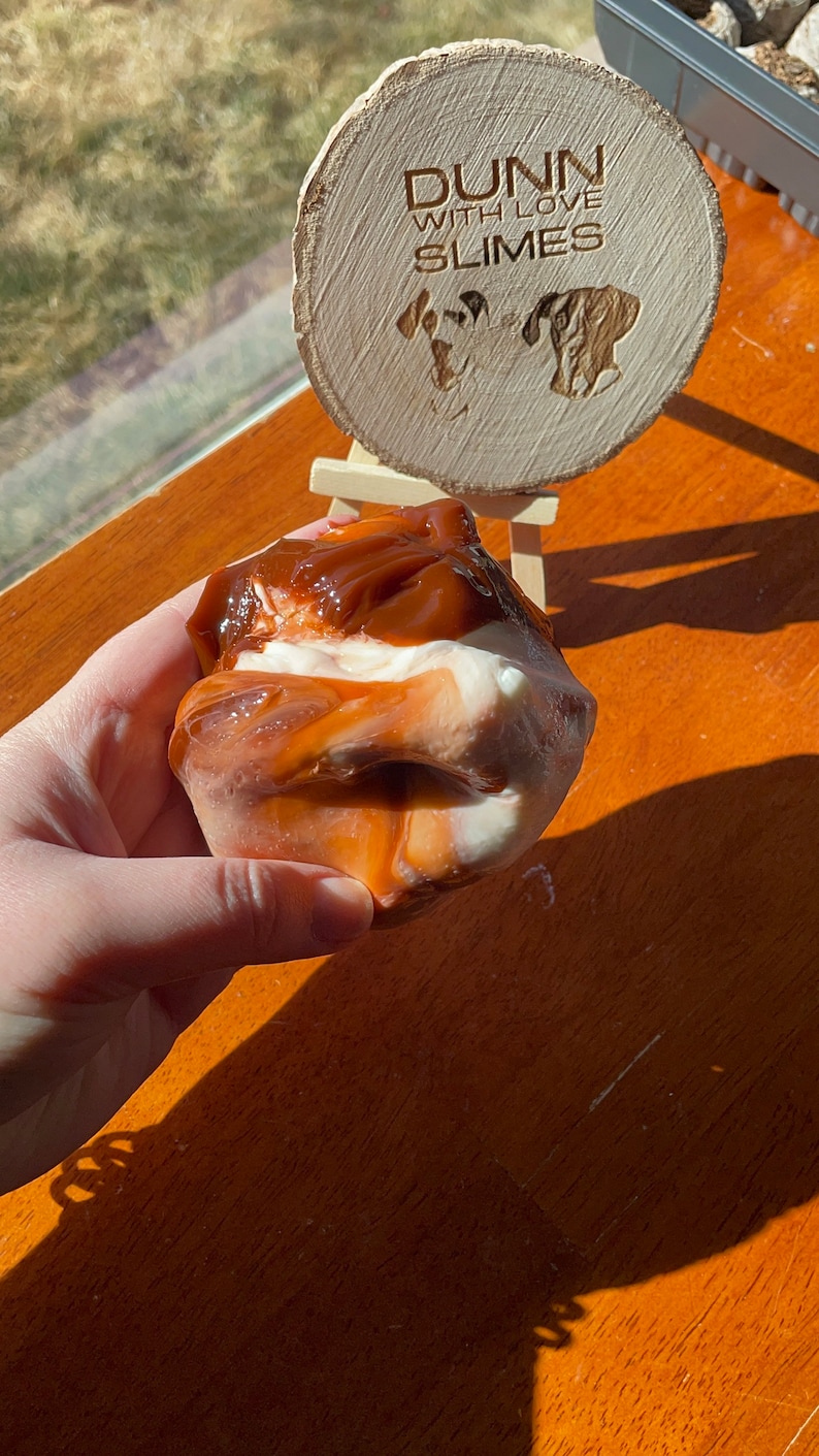 May include: A close-up of a brown and white slime with a caramel-colored swirl. The slime is being held in a hand. A wooden sign with the text "Dunn with Love Slimes" is in the background.