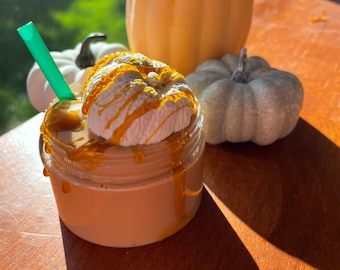 May include: A glass jar filled with a brown liquid topped with whipped cream and drizzled with caramel sauce. A green straw sticks out of the jar. The jar is sitting on a wooden table with two pumpkins in the background.