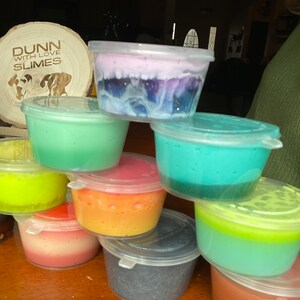 May include: A collection of colorful slime in clear plastic containers. The slime is in various colors, including green, blue, yellow, orange, red, and purple. The containers are stacked on top of each other. The slime is a popular toy for children.