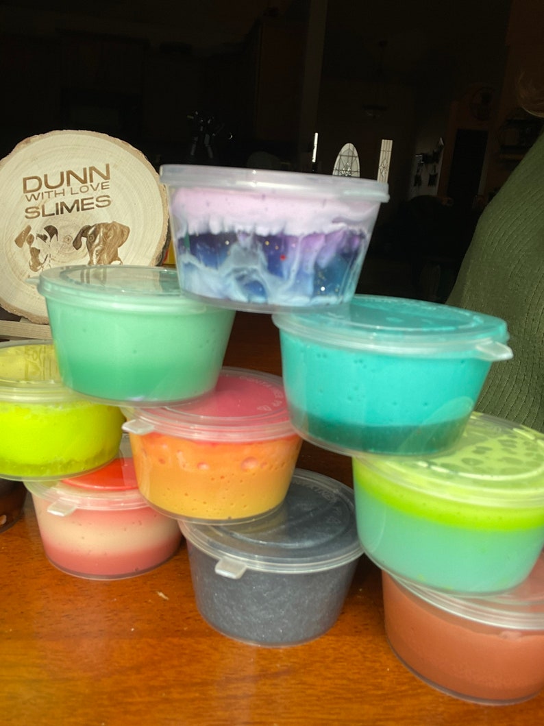 May include: A collection of colorful slime in clear plastic containers. The slime is in various colors, including green, blue, yellow, orange, red, and purple. The containers are stacked on top of each other. The slime is a popular toy for children.