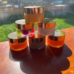 May include: Six small glass jars with gold lids, filled with different colored creams and gels. The jars are arranged in a pyramid shape on a wooden surface.
