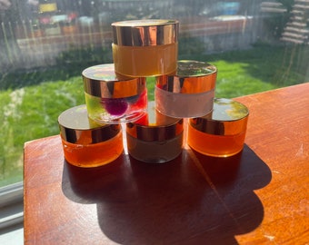 May include: Six small glass jars with gold lids, filled with different colored creams and gels. The jars are arranged in a pyramid shape on a wooden surface.