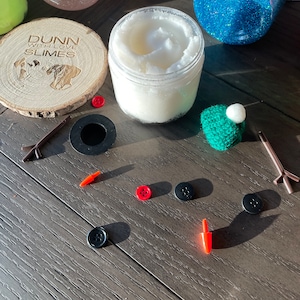 May include: A snowman craft kit with a clear jar of white slime, a green felt hat, black buttons, red buttons, and brown sticks. The kit also includes a wooden circle with the text "Dunn with Love Slimes".
