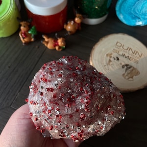 May include: A clear slime with red and silver glitter. The slime is in a hand and is being held up for the camera.