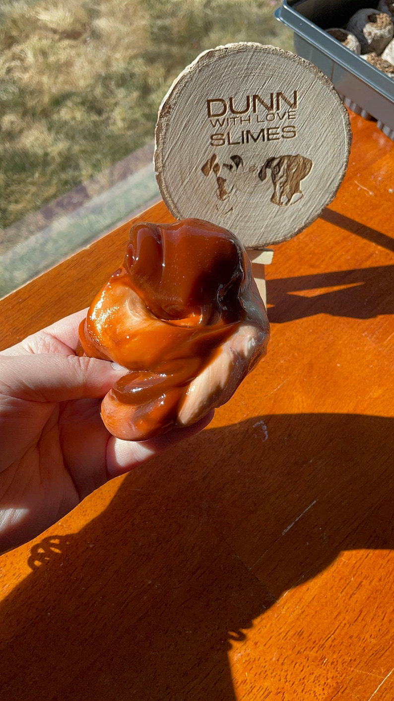 May include: A brown and white slime with a swirl pattern. The slime is being held in a hand. The slime is on a wooden surface. The slime is labeled "Dunn with Love Slimes".