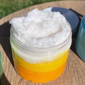 May include: A clear glass jar filled with a layered, colorful product resembling candy corn. The layers are orange, yellow, and white, topped with a fluffy white substance. The jar is sitting on a wooden surface.