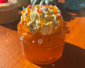 May include: A clear glass jar filled with a golden orange slime-like substance topped with a white whipped cream-like substance with caramel drizzle and colourful sprinkles.