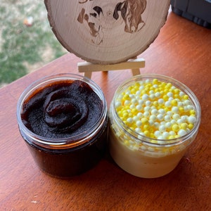 May include: Two glass jars of slime. One jar is filled with dark brown slime and the other jar is filled with light yellow slime with white beads. The jars are sitting on a wooden surface. The sign above the jars reads "Dunk with Love Slimes."