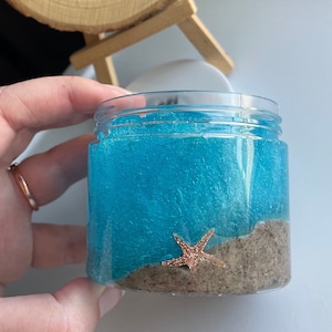 May include: A clear glass jar filled with blue slime with a layer of brown sand at the bottom. A gold starfish is partially submerged in the sand.