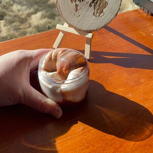 May include: A clear plastic jar filled with a brown and white slime. The slime is shaped like a swirl. The jar is sitting on a wooden table. A wooden sign with the text "Dunn with Love Slimes" is in the background.