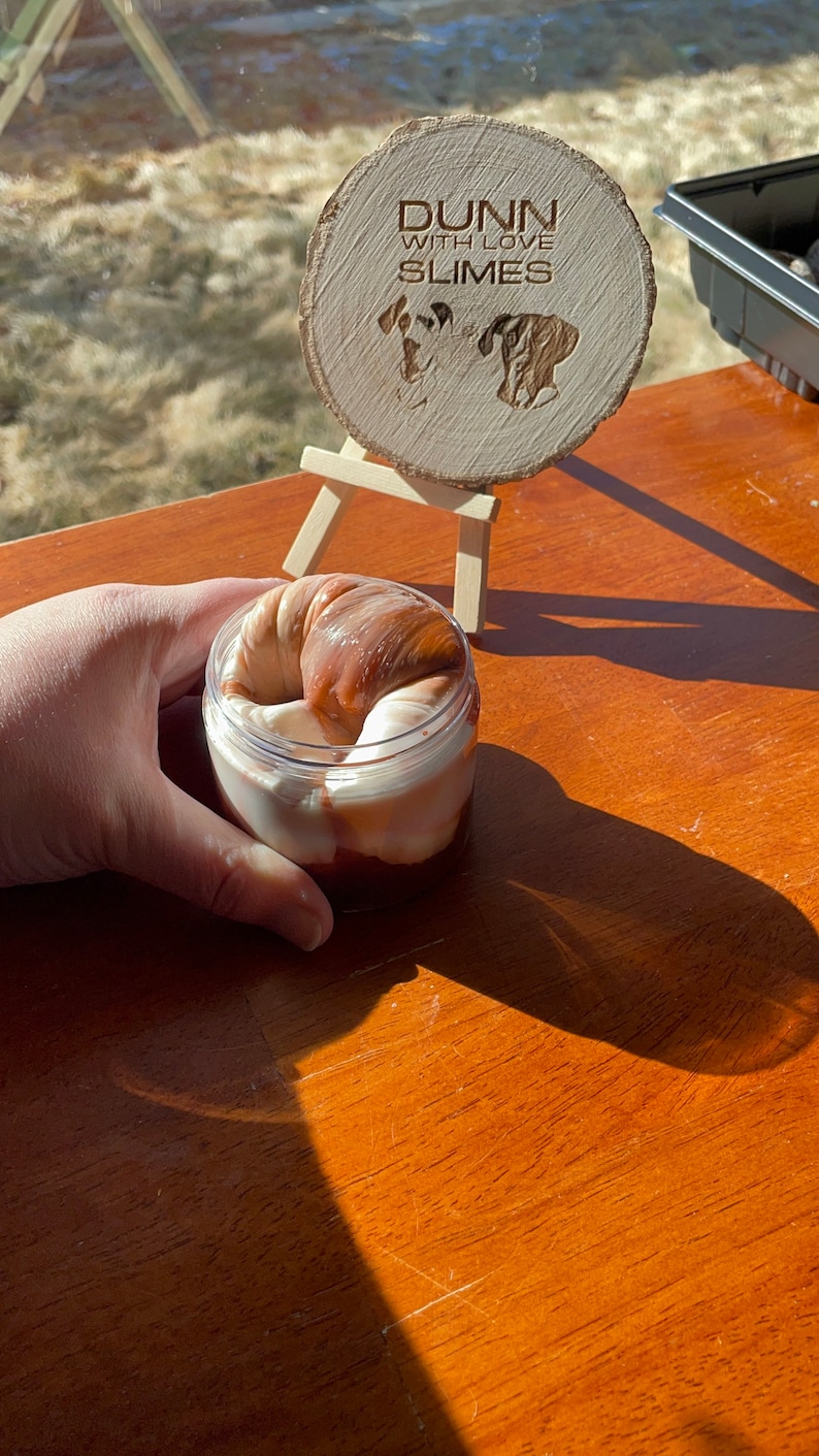 May include: A clear plastic jar filled with a brown and white slime. The slime is shaped like a swirl. The jar is sitting on a wooden table. A wooden sign with the text "Dunn with Love Slimes" is in the background.