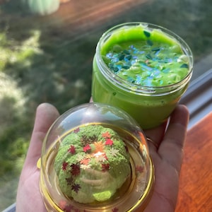 May include: A clear plastic jar filled with bright green slime containing iridescent glitter, accompanied by a separate clear dome lid featuring a textured green pumpkin figurine decorated with small metallic red and gold leaf-shaped sequins.