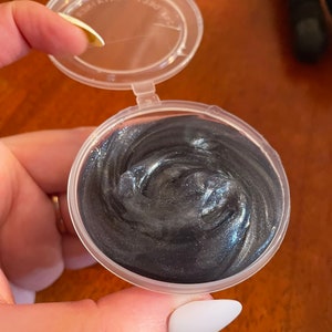 May include: A small clear plastic container with a lid, filled with a dark gray, shimmery slime.