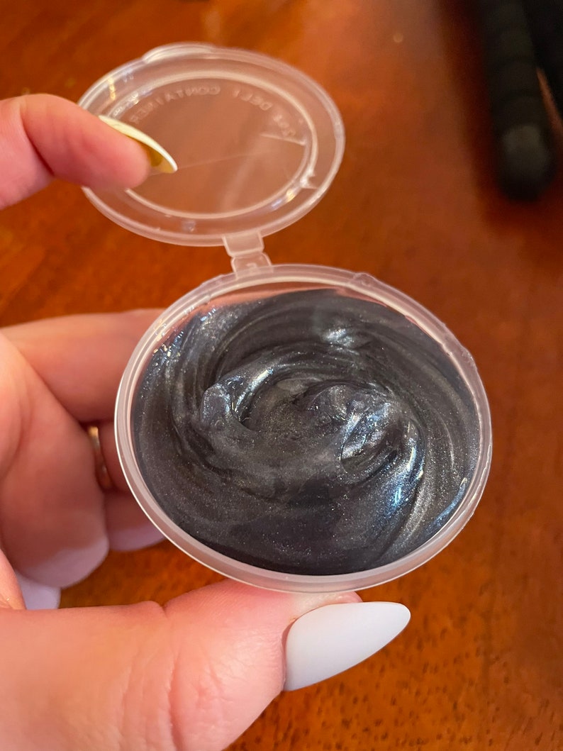 May include: A small clear plastic container with a lid, filled with a dark gray, shimmery slime.