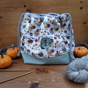Könnte beinhalten: Ein Kordelzugbeutel aus weißem Stoff mit einem Halloween-Cupcake-Muster, inklusive Fledermäusen und Wimpelketten. Der Boden ist einfarbig hellblau und in der Mitte befindet sich ein runder Aufnäher mit dem Text Handknit Hero.